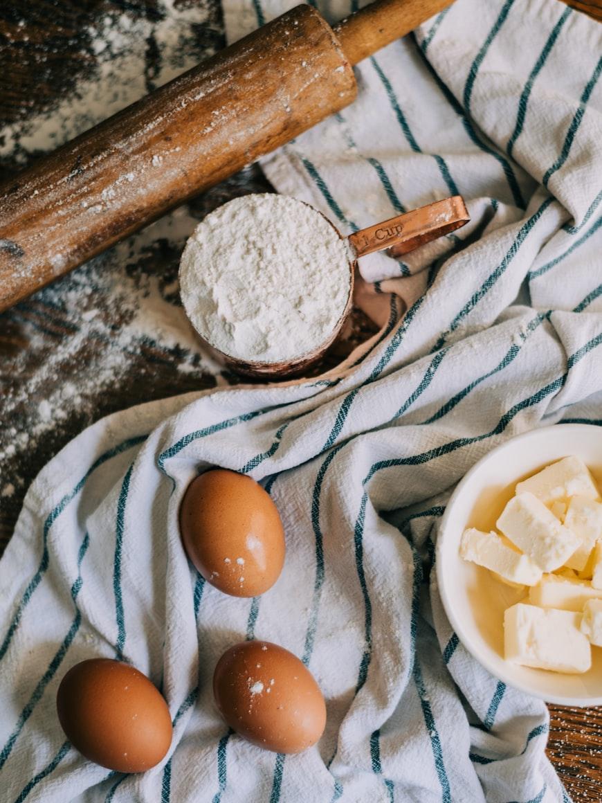 A flour-dusted rolling pin beside a copper measuring cup of flour, brown eggs and butter on a striped cloth