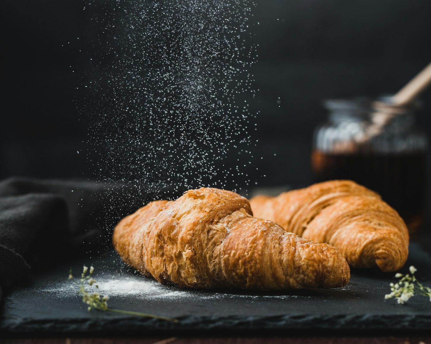 Two golden croissants on a slate board with icing sugar drifting down through the air