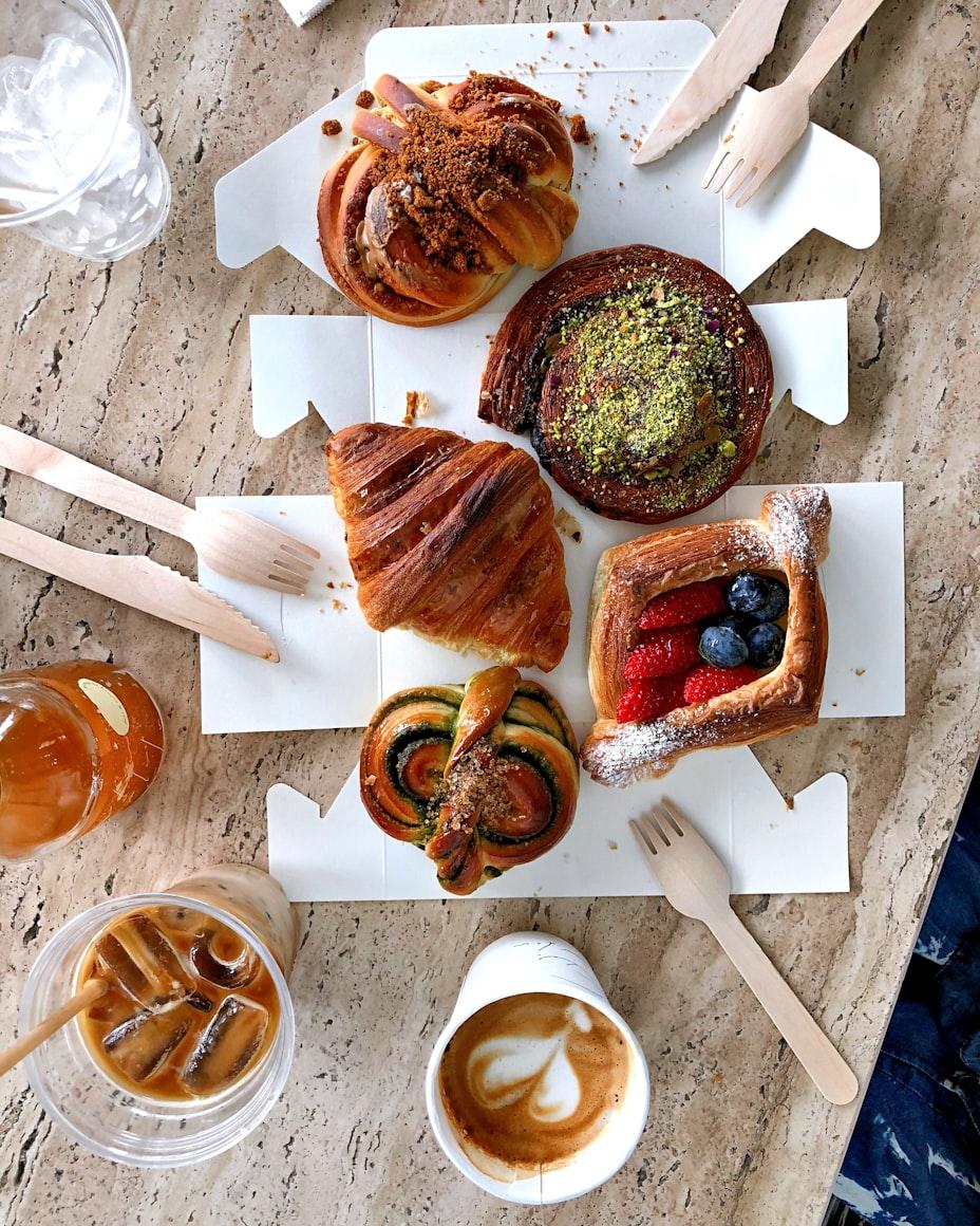 An assortment of fresh pastries on takeaway boards beside an iced coffee and a latte