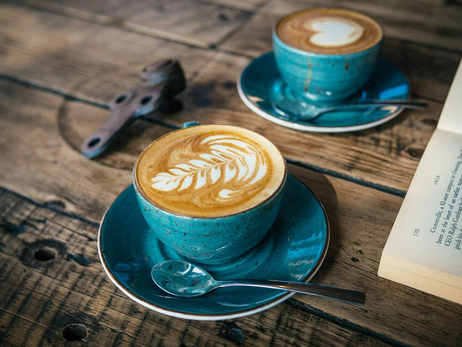 Two cappuccinos in teal speckled cups on a rustic wooden table with a vintage book