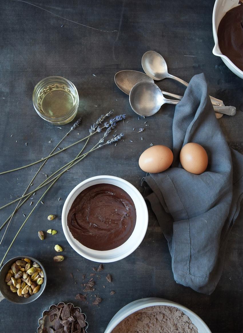 A bowl of glossy chocolate ganache surrounded by spoons, eggs and pistachios on a dark surface