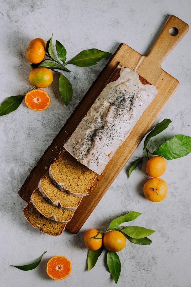 A glazed citrus poppyseed cake, partly sliced, on a wooden board with mandarins and leaves